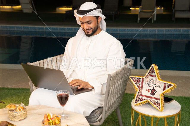 ضيافة شهر رمضان المبارك، تعابير السرور والسعادة، استخدام جهاز تقني حديث، شاب عربي خليجي اماراتي يرتدي الكندورة والغترة يعمل على الحاسب المحمول يجلس في الهواء الطلق، زينة واضاءة رمضانية، الجلسة الليلة الخارجية في احدى شاليهات الامارات