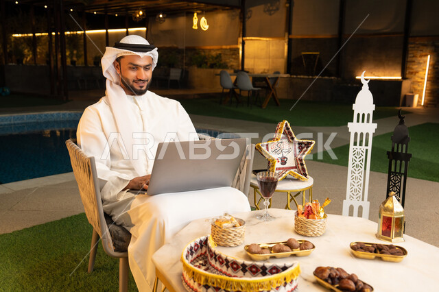 ضيافة شهر رمضان المبارك، تعابير السرور والسعادة، استخدام جهاز تقني حديث، شاب عربي خليجي اماراتي يرتدي الكندورة والغترة يعمل على الحاسب المحمول يجلس في الهواء الطلق، زينة واضاءة رمضانية، الجلسة الليلة الخارجية في احدى شاليهات الامارات