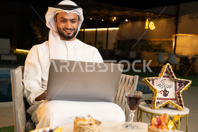 ضيافة شهر رمضان المبارك، تعابير السرور والسعادة، استخدام جهاز تقني حديث، شاب عربي خليجي اماراتي يرتدي الكندورة والغترة يعمل على الحاسب المحمول يجلس في الهواء الطلق، زينة واضاءة رمضانية، الجلسة الليلة الخارجية في احدى شاليهات الامارات