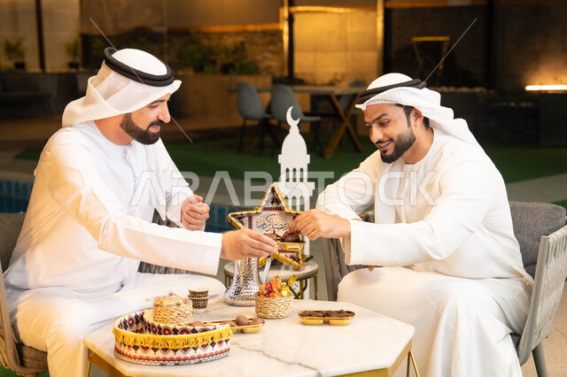 Eating fresh dates, a local Emirati product, Ramadan decorations and lighting, an outdoor evening session in one of the Emirati chalets, warm welcome and generosity in the holy month of Ramadan, two Emirati Gulf Arab friends wearing kanduras and ghutras sitting outdoors