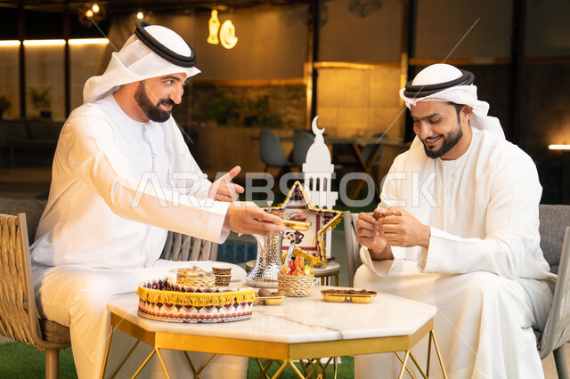 تناول التمر السكري الطازج، منتج وطني محلي اماراتي، زينة واضاءة رمضانية، الجلسة الليلة الخارجية في احدى شاليهات الامارات، حسن الاستقبال والكرم في شهر رمضان المبارك، صديقان عربيان خليجيان اماراتيان يرتديان الكندورة والغترة يجلسان في الهواء الطلق