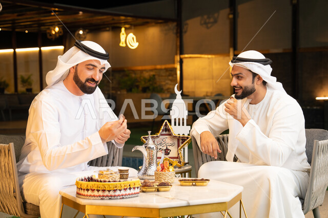 تناول التمر السكري الطازج، منتج وطني محلي اماراتي، زينة واضاءة رمضانية، الجلسة الليلة الخارجية في احدى شاليهات الامارات، حسن الاستقبال والكرم في شهر رمضان المبارك، صديقان عربيان خليجيان اماراتيان يرتديان الكندورة والغترة يجلسان في الهواء الطلق