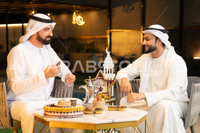 تناول التمر السكري الطازج، منتج وطني محلي اماراتي، زينة واضاءة رمضانية، الجلسة الليلة الخارجية في احدى شاليهات الامارات، حسن الاستقبال والكرم في شهر رمضان المبارك، صديقان عربيان خليجيان اماراتيان يرتديان الكندورة والغترة يجلسان في الهواء الطلق