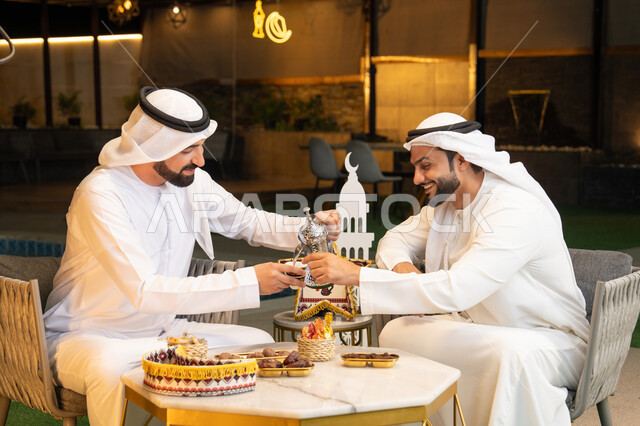 عادات وتقاليد شرب القهوة التقليدية، الجلسة الليلة الخارجية في احدى شاليهات الامارات، حسن الاستقبال والكرم في شهر رمضان المبارك، صديقان عربيان خليجيان اماراتيان يرتديان الكندورة والغترة يجلسان في الهواء الطلق، منتج وطني محلي اماراتي، زينة واضاءة رمضانية