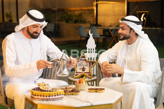 منتج وطني محلي اماراتي، زينة واضاءة رمضانية، عادات وتقاليد شرب القهوة التقليدية، الجلسة الليلة الخارجية في احدى شاليهات الامارات، حسن الاستقبال والكرم في شهر رمضان المبارك، صديقان عربيان خليجيان اماراتيان يرتديان الكندورة والغترة يجلسان في الهواء الطلق