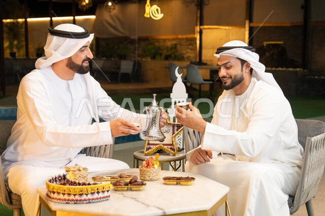منتج وطني محلي اماراتي، زينة واضاءة رمضانية، عادات وتقاليد شرب القهوة التقليدية، الجلسة الليلة الخارجية في احدى شاليهات الامارات، حسن الاستقبال والكرم في شهر رمضان المبارك، صديقان عربيان خليجيان اماراتيان يرتديان الكندورة والغترة يجلسان في الهواء الطلق