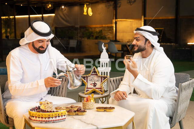 عادات وتقاليد شرب القهوة التقليدية، الجلسة الليلة الخارجية في احدى شاليهات الامارات، حسن الاستقبال والكرم في شهر رمضان المبارك، صديقان عربيان خليجيان اماراتيان يرتديان الكندورة والغترة يجلسان في الهواء الطلق، منتج وطني محلي اماراتي، زينة واضاءة رمضانية