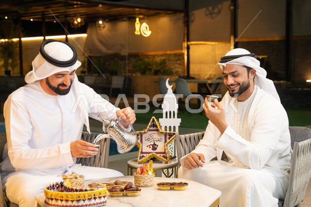 منتج وطني محلي اماراتي، زينة واضاءة رمضانية، عادات وتقاليد شرب القهوة التقليدية، الجلسة الليلة الخارجية في احدى شاليهات الامارات، حسن الاستقبال والكرم في شهر رمضان المبارك، صديقان عربيان خليجيان اماراتيان يرتديان الكندورة والغترة يجلسان في الهواء الطلق
