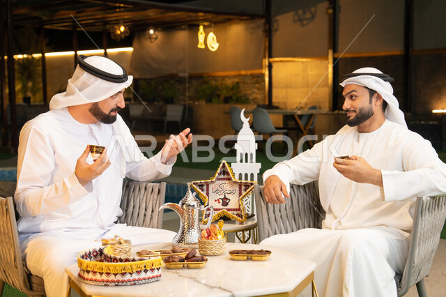 تبادل الاحاديث والاخبار بتعابير السرور، زينة واضاءة رمضانية، الجلسة الليلة الخارجية في احدى شاليهات الامارات، حسن الاستقبال وكرم الضيافة في شهر رمضان المبارك، صديقان عربيان خليجيان اماراتيان يرتديان الكندورة والغترة يجلسان في الهواء الطلق