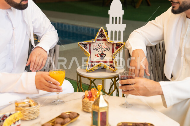 An outdoor evening gathering in one of the chalets in the Emirates, warm welcome and generosity, a close-up of the hospitality table in the holy month of Ramadan, two Emirati Gulf Arab friends wearing kanduras and ghutras sitting in the open air, exchanging conversations and news with expressions of pleasure, Ramadan decorations and lighting