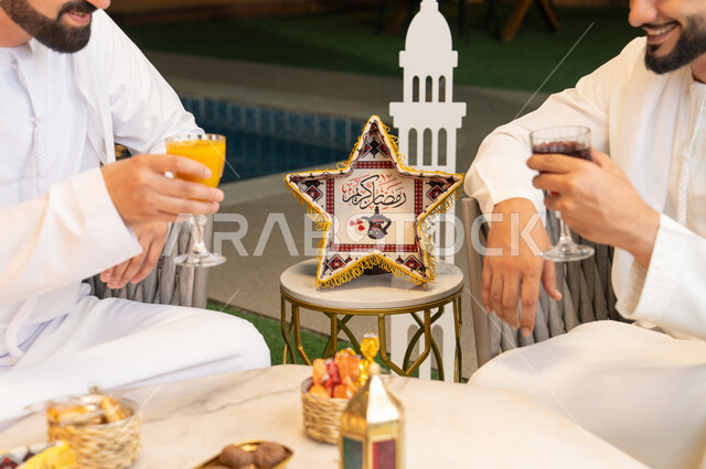الجلسة الليلة الخارجية في احدى شاليهات الامارات، حسن الاستقبال والكرم، صورة مقربة لطاولة الضيافة في شهر رمضان المبارك، صديقان عربيان خليجيان اماراتيان يرتديان الكندورة والغترة يجلسان في الهواء الطلق، تبادل الاحاديث والاخبار بتعابير السرور، زينة واضاءة رمضانية