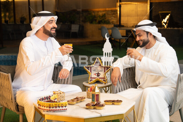 زينة واضاءة رمضانية، الجلسة الليلة الخارجية في احدى شاليهات الامارات، حسن الاستقبال وكرم الضيافة في شهر رمضان المبارك، صديقان عربيان خليجيان اماراتيان يرتديان الكندورة والغترة يجلسان في الهواء الطلق، تبادل الاحاديث والاخبار بتعابير السرور