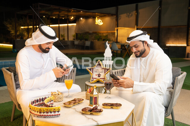 Using a mobile phone with expressions of integration, Ramadan decorations and lighting, an outdoor evening session in one of the chalets in the Emirates, warm welcome and hospitality during the holy month of Ramadan, two Emirati Gulf Arab friends wearing kanduras and ghutras sitting outdoors
