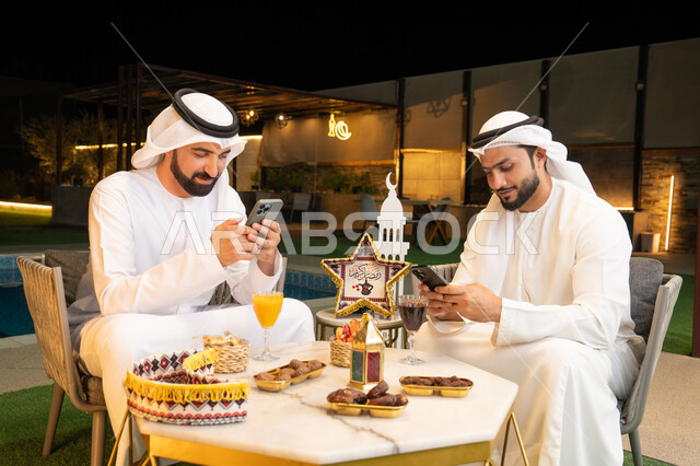 استخدام الهاتف المحمول بتعابير الاندماج، زينة واضاءة رمضانية، الجلسة الليلة الخارجية في احدى شاليهات الامارات، حسن الاستقبال وكرم الضيافة في شهر رمضان المبارك، صديقان عربيان خليجيان اماراتيان يرتديان الكندورة والغترة يجلسان في الهواء الطلق