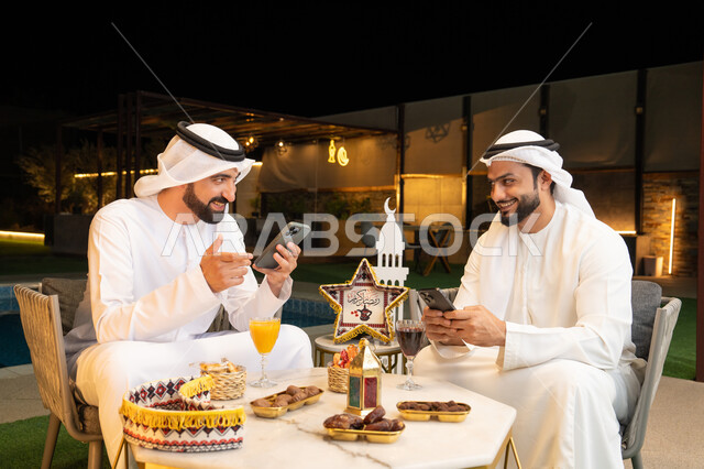 استخدام الهاتف المحمول بتعابير الاندماج، زينة واضاءة رمضانية، الجلسة الليلة الخارجية في احدى شاليهات الامارات، حسن الاستقبال وكرم الضيافة في شهر رمضان المبارك، صديقان عربيان خليجيان اماراتيان يرتديان الكندورة والغترة يجلسان في الهواء الطلق
