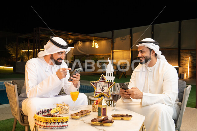 استخدام الهاتف المحمول بتعابير الاندماج، زينة واضاءة رمضانية، الجلسة الليلة الخارجية في احدى شاليهات الامارات، حسن الاستقبال وكرم الضيافة في شهر رمضان المبارك، صديقان عربيان خليجيان اماراتيان يرتديان الكندورة والغترة يجلسان في الهواء الطلق