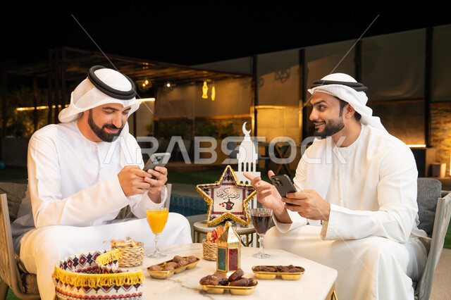 استخدام الهاتف المحمول بتعابير الاندماج، زينة واضاءة رمضانية، الجلسة الليلة الخارجية في احدى شاليهات الامارات، حسن الاستقبال وكرم الضيافة في شهر رمضان المبارك، صديقان عربيان خليجيان اماراتيان يرتديان الكندورة والغترة يجلسان في الهواء الطلق