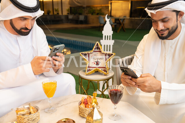 استخدام الهاتف المحمول بتعابير الاندماج، زينة واضاءة رمضانية، الجلسة الليلة الخارجية في احدى شاليهات الامارات، حسن الاستقبال وكرم الضيافة في شهر رمضان المبارك، صديقان عربيان خليجيان اماراتيان يرتديان الكندورة والغترة يجلسان في الهواء الطلق