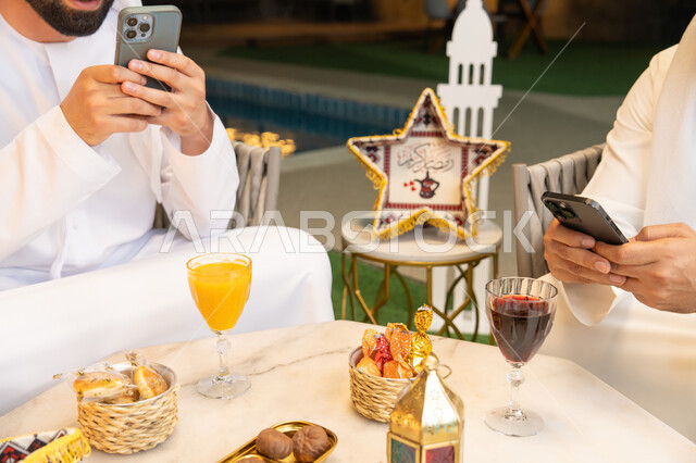 استخدام الهاتف المحمول بتعابير الاندماج، زينة واضاءة رمضانية، الجلسة الليلة الخارجية في احدى شاليهات الامارات، حسن الاستقبال وكرم الضيافة في شهر رمضان المبارك، صديقان عربيان خليجيان اماراتيان يرتديان الكندورة والغترة يجلسان في الهواء الطلق