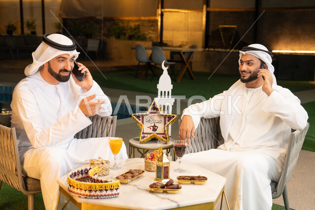 اجراء مكالمة هاتفية باستخدام الهاتف المحمول بتعابير السرور، زينة واضاءة رمضانية، الجلسة الليلة الخارجية في احدى شاليهات الامارات، حسن الاستقبال وكرم الضيافة في شهر رمضان المبارك، صديقان عربيان خليجيان اماراتيان يرتديان الكندورة والغترة يجلسان في الهواء الطلق