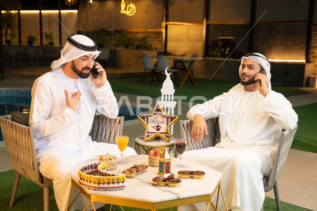 اجراء مكالمة هاتفية باستخدام الهاتف المحمول بتعابير السرور، زينة واضاءة رمضانية، الجلسة الليلة الخارجية في احدى شاليهات الامارات، حسن الاستقبال وكرم الضيافة في شهر رمضان المبارك، صديقان عربيان خليجيان اماراتيان يرتديان الكندورة والغترة يجلسان في الهواء الطلق