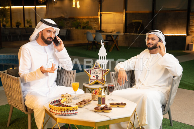 اجراء مكالمة هاتفية باستخدام الهاتف المحمول بتعابير السرور، زينة واضاءة رمضانية، الجلسة الليلة الخارجية في احدى شاليهات الامارات، حسن الاستقبال وكرم الضيافة في شهر رمضان المبارك، صديقان عربيان خليجيان اماراتيان يرتديان الكندورة والغترة يجلسان في الهواء الطلق