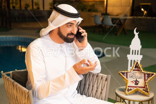 الرد على الاتصال الهاتفي بتعابير السرور، ضيافة شهر رمضان المبارك، استخدام جهاز تقني حديث، شاب عربي خليجي اماراتي يرتدي الكندورة والغترة يجري مكالمة هاتفية عبر الهاتف المحمول يجلس في الهواء الطلق، زينة واضاءة رمضانية، الجلسة الليلة الخارجية في احدى الشاليهات