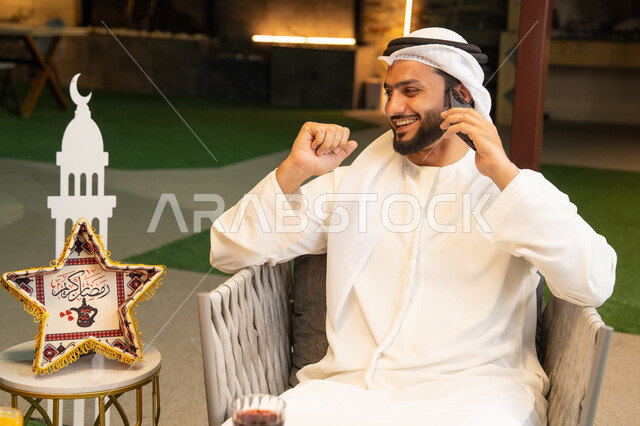 الرد على الاتصال الهاتفي بتعابير السرور، ضيافة شهر رمضان المبارك، استخدام جهاز تقني حديث، شاب عربي خليجي اماراتي يرتدي الكندورة والغترة يجري مكالمة هاتفية عبر الهاتف المحمول يجلس في الهواء الطلق، زينة واضاءة رمضانية، الجلسة الليلة الخارجية في احدى الشاليهات
