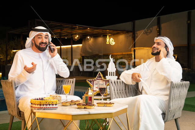 Making a phone call using a mobile phone with happy expressions, Ramadan decorations and lighting, sitting outside at a chalet in the Emirates at night, warm welcome and hospitality during the holy month of Ramadan, two Emirati Gulf Arab friends wearing kandura and ghutra sitting outdoors