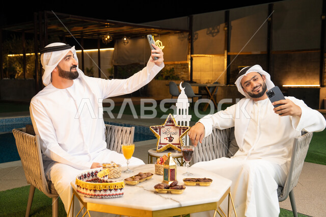 توثيق اللحظات السعيدة، حسن الاستقبال وكرم الضيافة في شهر رمضان المبارك، صديقان عربيان خليجيان اماراتيان يرتديان الكندورة والغترة يجلسان في الهواء الطلق، التقاط صور سيلفي  باستخدام الهاتف المحمول بتعابير السرور، زينة واضاءة رمضانية، الجلسة الليلة الخارجية
