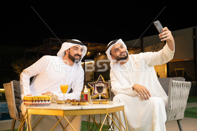 التقاط صور سيلفي  باستخدام الهاتف المحمول بتعابير السرور، زينة واضاءة رمضانية، الجلسة الليلة الخارجية، توثيق اللحظات السعيدة، حسن الاستقبال وكرم الضيافة في شهر رمضان المبارك، صديقان عربيان خليجيان اماراتيان يرتديان الكندورة والغترة يجلسان في الهواء الطلق