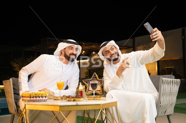 Documenting happy moments, warm welcome and hospitality during the holy month of Ramadan, two Emirati Gulf Arab friends wearing kandura and ghutra sitting outdoors, taking selfies using mobile phones with happy expressions, Ramadan decorations and lighting, outdoor night session