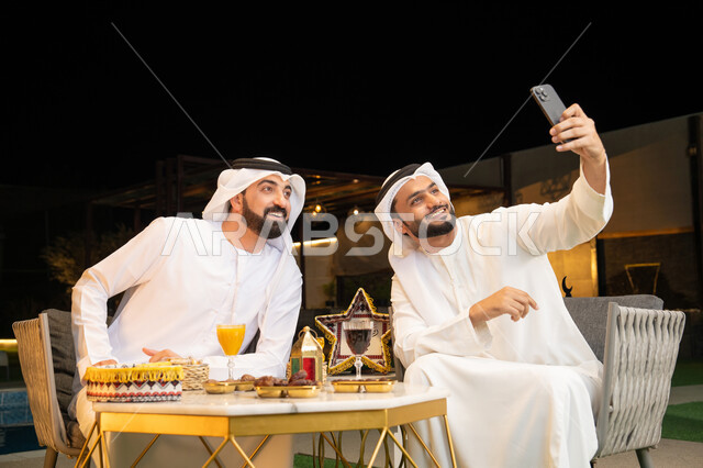 Documenting happy moments, warm welcome and hospitality during the holy month of Ramadan, two Emirati Gulf Arab friends wearing kandura and ghutra sitting outdoors, taking selfies using mobile phones with happy expressions, Ramadan decorations and lighting, outdoor night session
