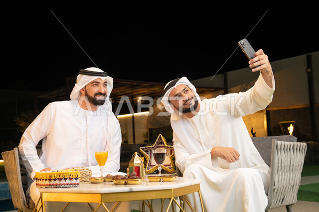 Documenting happy moments, warm welcome and hospitality during the holy month of Ramadan, two Emirati Gulf Arab friends wearing kandura and ghutra sitting outdoors, taking selfies using mobile phones with happy expressions, Ramadan decorations and lighting, outdoor night session
