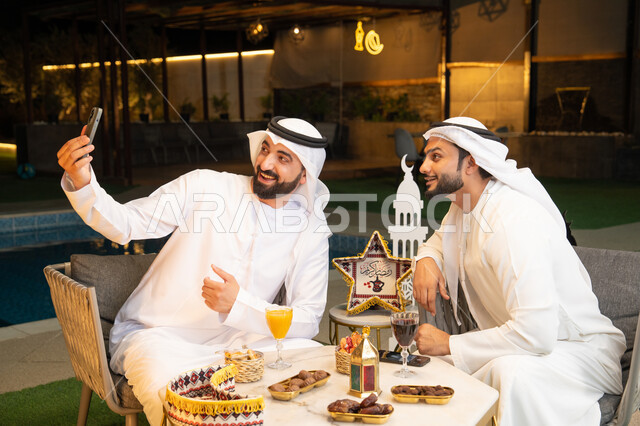 Documenting happy moments, warm welcome and hospitality during the holy month of Ramadan, two Emirati Gulf Arab friends wearing kandura and ghutra sitting outdoors, taking selfies using mobile phones with happy expressions, Ramadan decorations and lighting, outdoor night session