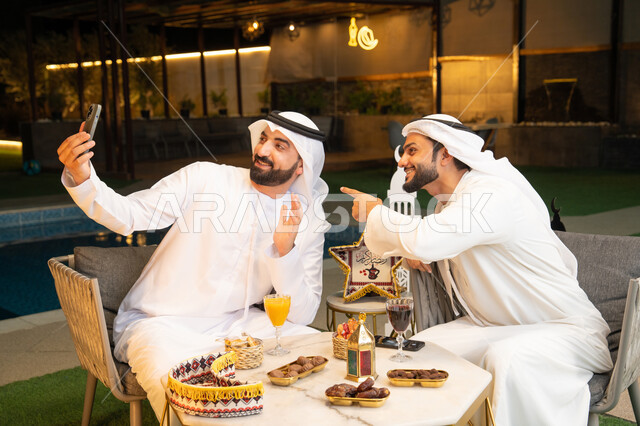 التقاط صور سيلفي  باستخدام الهاتف المحمول بتعابير السرور، زينة واضاءة رمضانية، الجلسة الليلة الخارجية، توثيق اللحظات السعيدة، حسن الاستقبال وكرم الضيافة في شهر رمضان المبارك، صديقان عربيان خليجيان اماراتيان يرتديان الكندورة والغترة يجلسان في الهواء الطلق