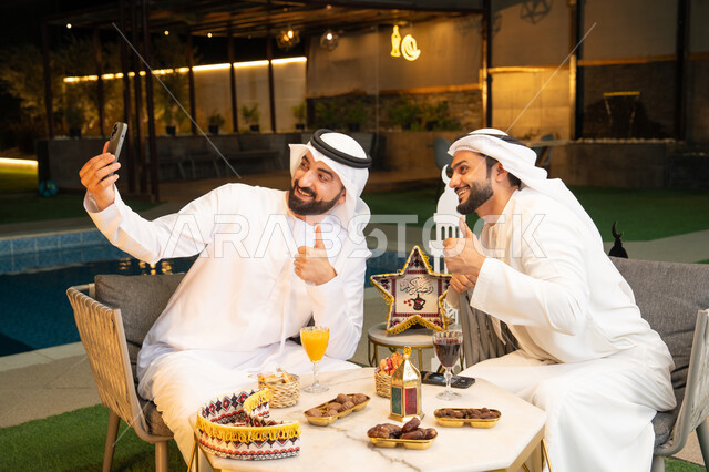 توثيق اللحظات السعيدة، حسن الاستقبال وكرم الضيافة في شهر رمضان المبارك، صديقان عربيان خليجيان اماراتيان يرتديان الكندورة والغترة يجلسان في الهواء الطلق، التقاط صور سيلفي  باستخدام الهاتف المحمول بتعابير السرور، زينة واضاءة رمضانية، الجلسة الليلة الخارجية