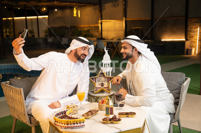 توثيق اللحظات السعيدة، حسن الاستقبال وكرم الضيافة في شهر رمضان المبارك، صديقان عربيان خليجيان اماراتيان يرتديان الكندورة والغترة يجلسان في الهواء الطلق، التقاط صور سيلفي  باستخدام الهاتف المحمول بتعابير السرور، زينة واضاءة رمضانية، الجلسة الليلة الخارجية