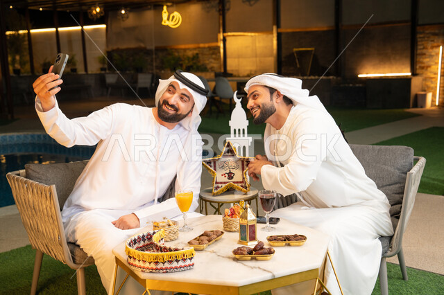 التقاط صور سيلفي  باستخدام الهاتف المحمول بتعابير السرور، زينة واضاءة رمضانية، الجلسة الليلة الخارجية، توثيق اللحظات السعيدة، حسن الاستقبال وكرم الضيافة في شهر رمضان المبارك، صديقان عربيان خليجيان اماراتيان يرتديان الكندورة والغترة يجلسان في الهواء الطلق