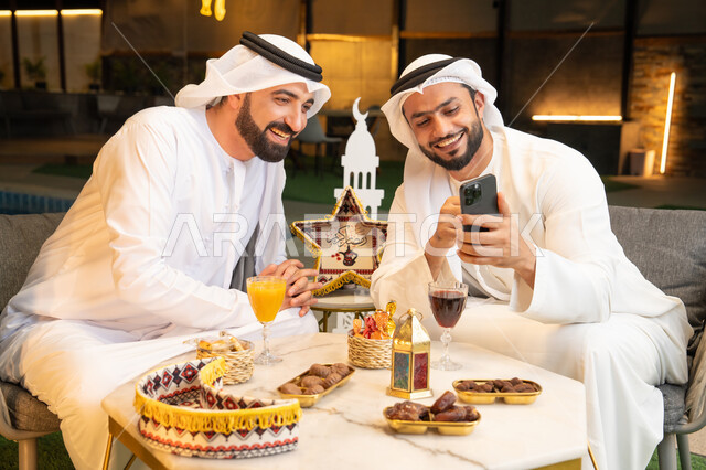 استخدام التقنية والهاتف المحمول بتعابير السرور والسعادة، زينة واضاءة رمضانية، الجلسة الليلة الخارجية في احدى شاليهات الامارات، حسن الاستقبال وكرم الضيافة في شهر رمضان المبارك، صديقان عربيان خليجيان اماراتيان يرتديان الكندورة والغترة يجلسان في الهواء الطلق