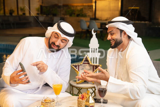 استخدام التقنية والهاتف المحمول بتعابير السرور والسعادة، زينة واضاءة رمضانية، الجلسة الليلة الخارجية في احدى شاليهات الامارات، حسن الاستقبال وكرم الضيافة في شهر رمضان المبارك، صديقان عربيان خليجيان اماراتيان يرتديان الكندورة والغترة يجلسان في الهواء الطلق