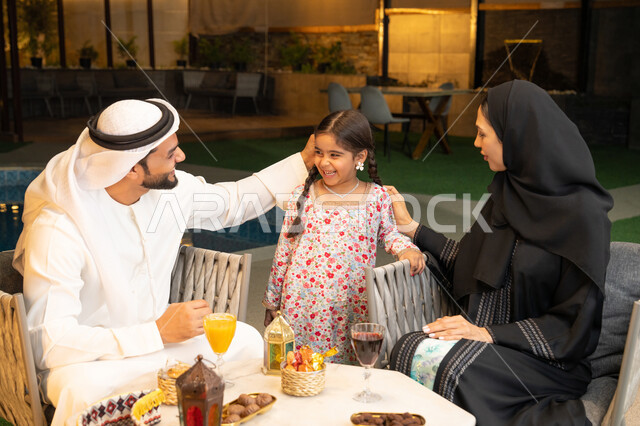 زينة واكسسوار الأعياد والمناسبات الاسلامية، فتاة عربية خليجية اماراتية تحمل بيدها فانوس معدني مزخرف مضيء بجانب والديها، قضاء وقت ممتع في جلسة خارجية عائلية في احدى الشاليهات، استقبال شهر رمضان المبارك، أجواء ليلية رمضانية روحانية