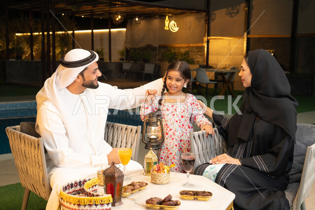 Spiritual Ramadan night atmosphere, decorations and accessories for Islamic holidays and occasions, an Emirati Gulf Arab girl holding a decorated illuminated metal lantern next to her parents, spending a pleasant time in an outdoor family session in one of the chalets, welcoming the holy month of Ramadan