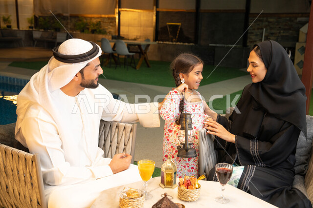 زينة واكسسوار الأعياد والمناسبات الاسلامية، فتاة عربية خليجية اماراتية تحمل بيدها فانوس معدني مزخرف مضيء بجانب والديها، قضاء وقت ممتع في جلسة خارجية عائلية في احدى الشاليهات، استقبال شهر رمضان المبارك، أجواء ليلية رمضانية روحانية