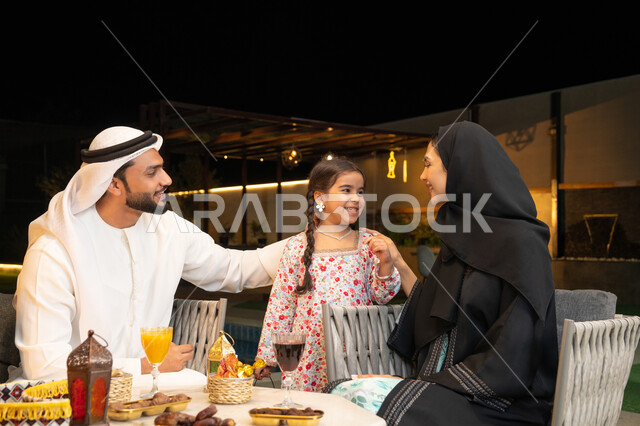 أجواء ليلية رمضانية روحانية، زينة واكسسوار الأعياد والمناسبات الاسلامية، فتاة عربية خليجية اماراتية تحمل بيدها فانوس معدني مزخرف مضيء بجانب والديها، قضاء وقت ممتع في جلسة خارجية عائلية في احدى الشاليهات، استقبال شهر رمضان المبارك