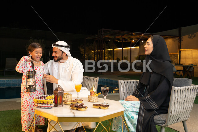 زينة واكسسوار الأعياد والمناسبات الاسلامية، فتاة عربية خليجية اماراتية تحمل بيدها فانوس معدني مزخرف مضيء بجانب والديها، قضاء وقت ممتع في جلسة خارجية عائلية في احدى الشاليهات، استقبال شهر رمضان المبارك، أجواء ليلية رمضانية روحانية