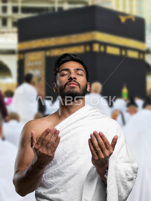 رفع اليدين والدعاء بخشوع وتضرع, أداء مناسك الحج و العمرة, أماكن و معالم دينة إسلامية مقدسة في المملكة العربية السعودية, العبادة و التقرب إلى الله, صورة مقربة لرجل عربي خليجي سعودي يرتدي ملابس الاحرام يقف امام الكعبه المشرفه رافعا يديه بالدعاء