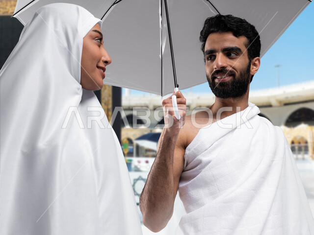 أداء مناسك الحج و العمرة, أماكن و معالم دينة إسلامية مقدسة في المملكة العربية السعودية, العبادة و التقرب إلى الله, صورة مقربة لزوجان عربيان خليجيان سعوديان مبتسمان يرتديان ملابس الاحرام يمسكان بمظلتهما الشمسيه استعدادا لاداء مناسك الحج