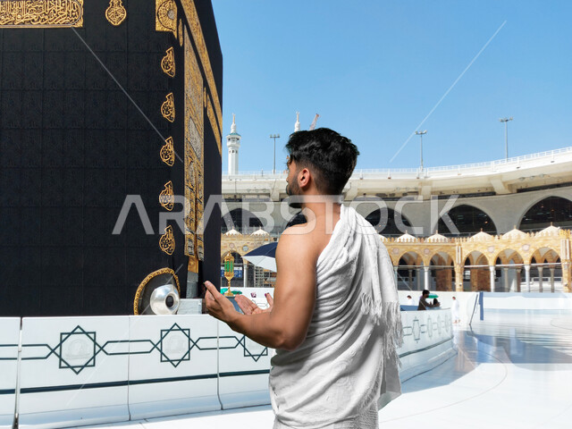العبادة و التقرب إلى الله, أداء مناسك الحج و العمرة, أماكن و معالم دينة إسلامية مقدسة في المملكة العربية السعودية, صورة من الخلف لرجل عربي خليجي سعودي يرتدي ملابس الاحرام يقف امام الكعبه الشريفه يقوم بالدعاء