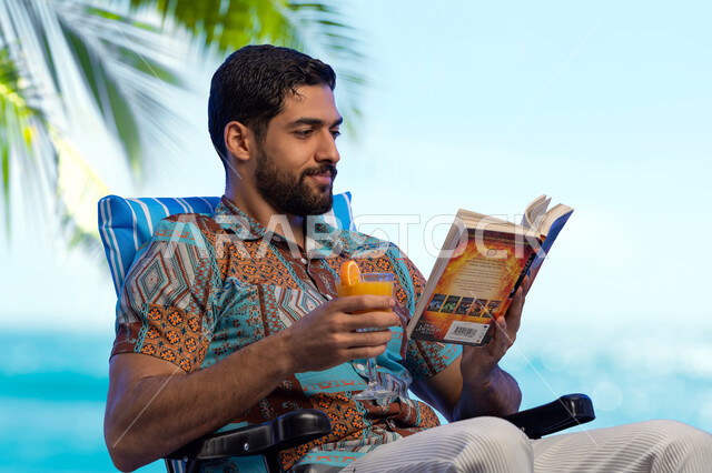 Sitting and enjoying the vacation and the scenery, practicing a useful hobby in free time, a smiling young Saudi Arabian Gulf man wearing casual clothes sitting on a comfortable chair with a happy expression reading a book, a pleasant youth trip, spending the summer vacation time on tourism